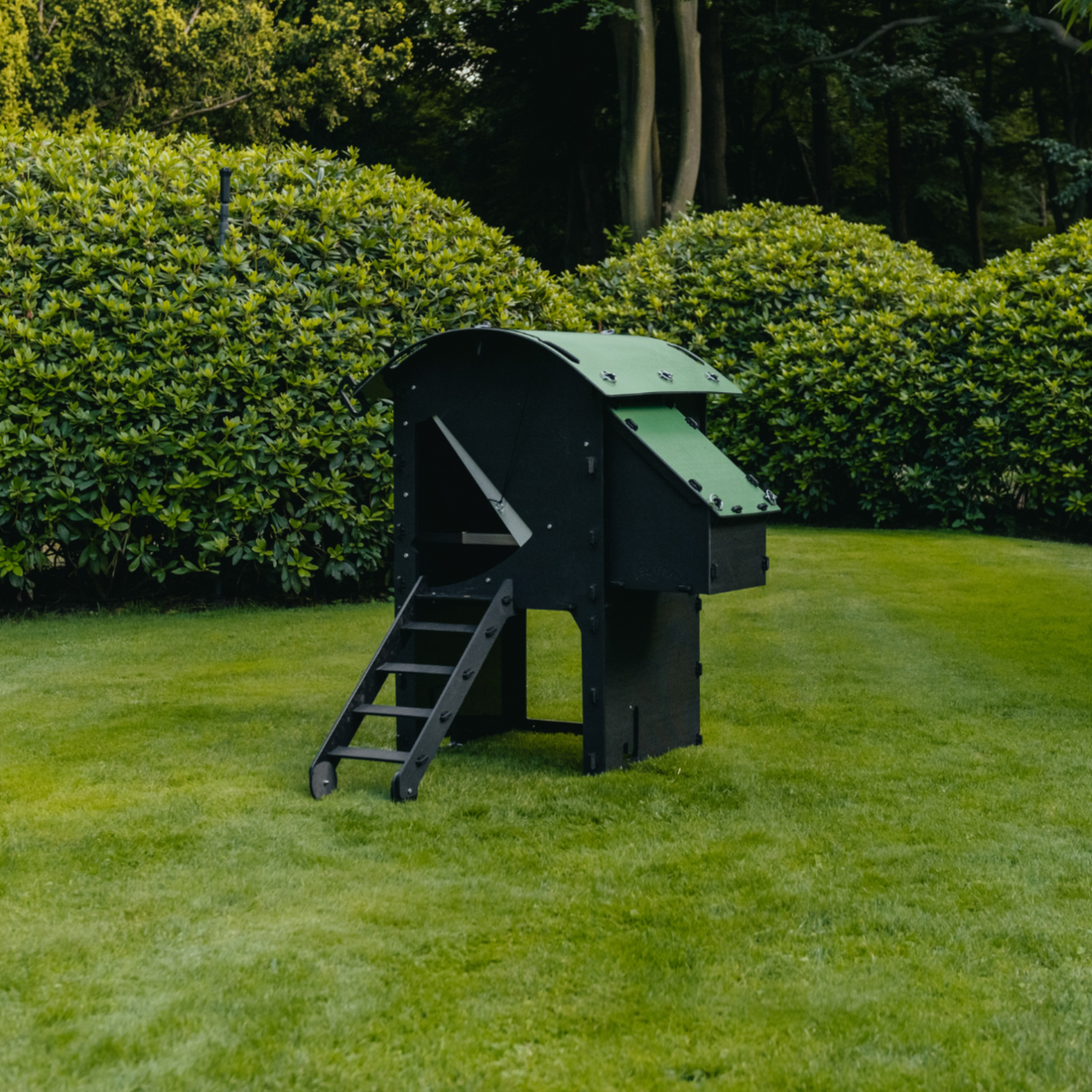 Hatching Time Nestera. Small Raised coop can be seen in image on grass. Coop has door open with automatic coop opener. Ramp is leading up to opening for easy chicken access. Nesting box is on side connected to chicken coop, with lifting door for easy egg access. Roof of coop is slanted to prevent roosting.