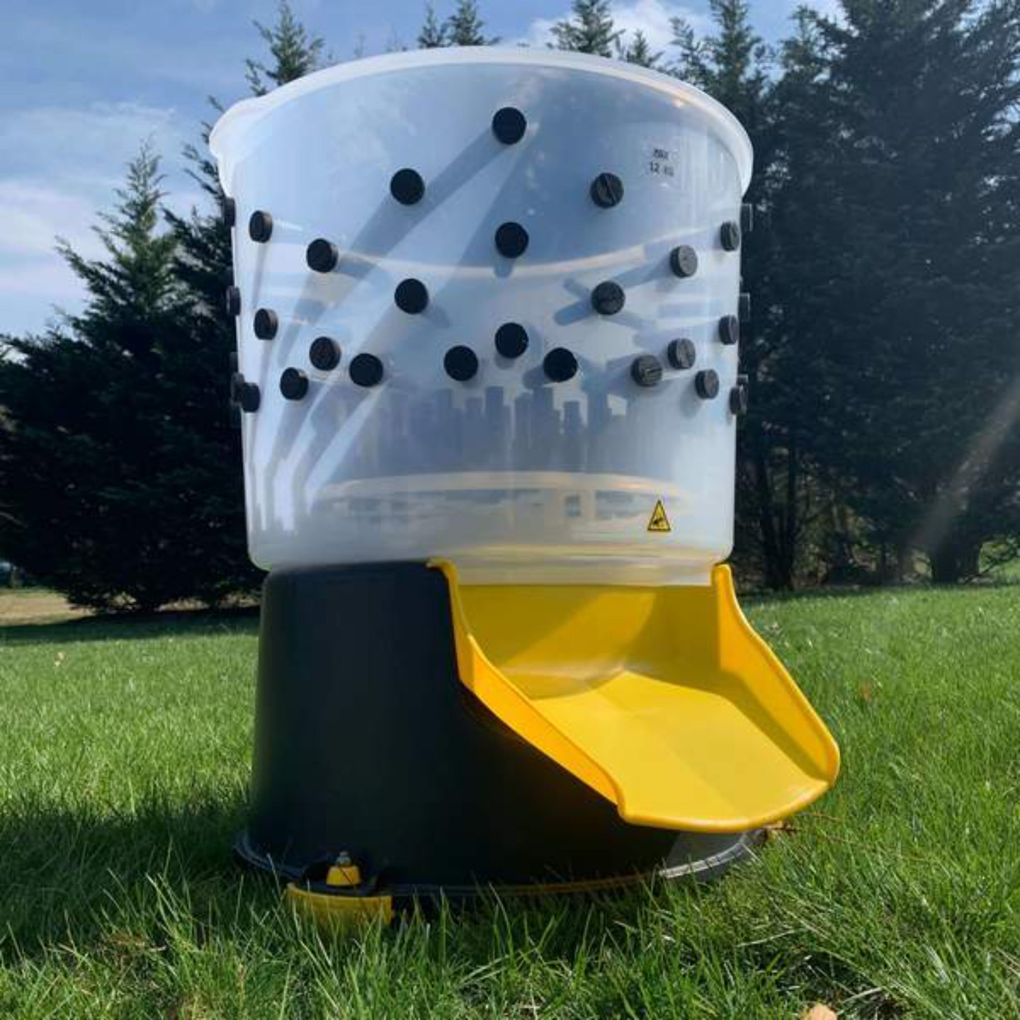 Hatching Time Cimuka. Goose feather plucker can be seen sitting on grass to show scale size of plucker. Feather chute is visible and all-natural rubber plucker fingers can be seen through rust-proof food grade plastic tub.