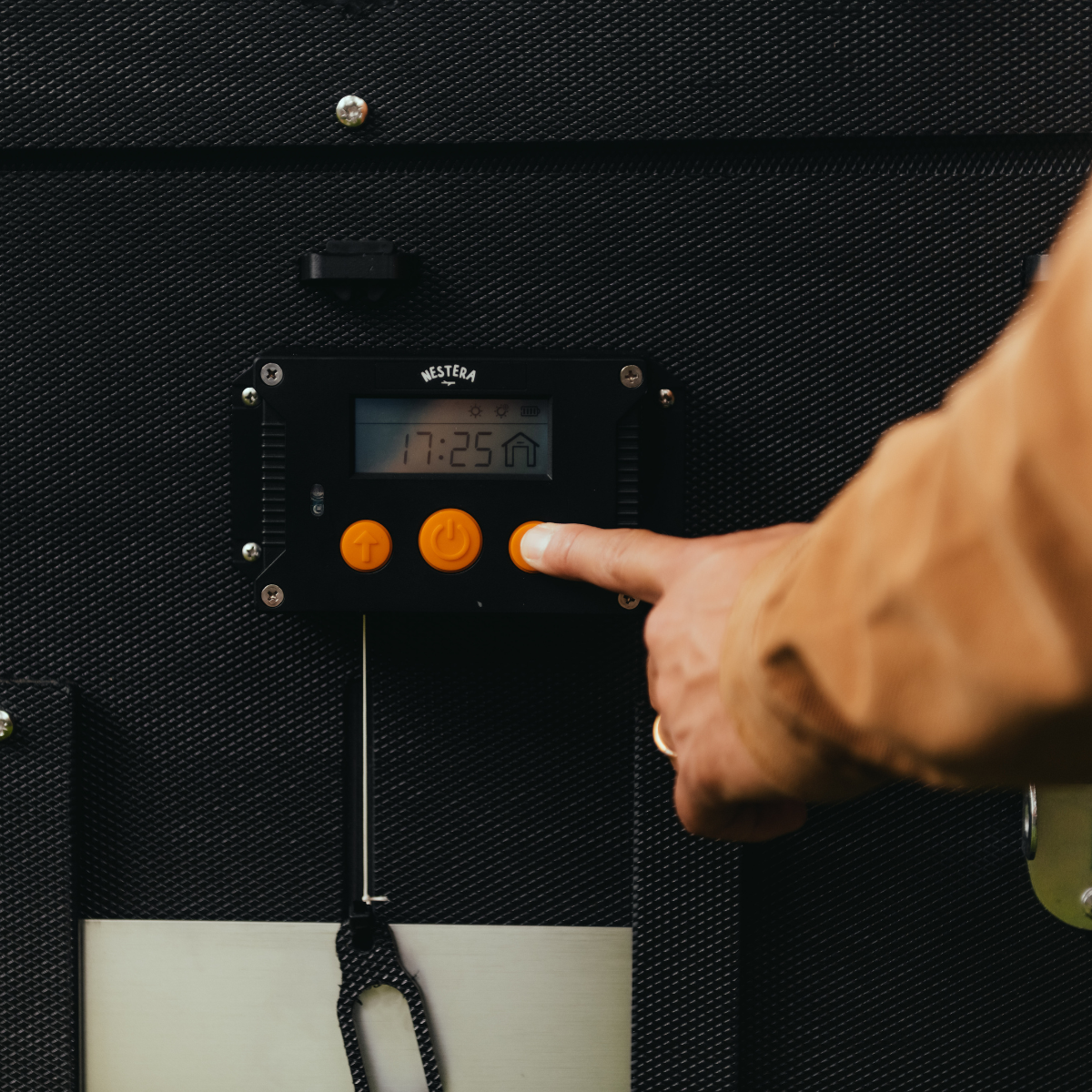 Hatching Time Nestera. A hand can be seen pressing the digital control for Automatic coop door opener that is mounted on exterior chicken coop wall.