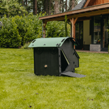 Hatching Time Nestera. Small coop can be seen in image on grass. Coop has door open with automatic coop opener. Ramp is leading up to opening for easy chicken access. Nesting box is on side connected to chicken coop, with lifting door for easy egg access. Roof of coop is slanted to prevent roosting.