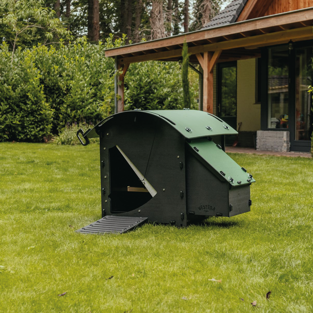 Hatching Time Nestera. Small ground coop can be seen in image in grass in front of house. Coop has door open with automatic coop opener. Ramp is leading up to opening for easy chicken access. Nesting box is on side connected to chicken coop, with lifting door for easy egg access. Roof of coop is slanted to prevent roosting.