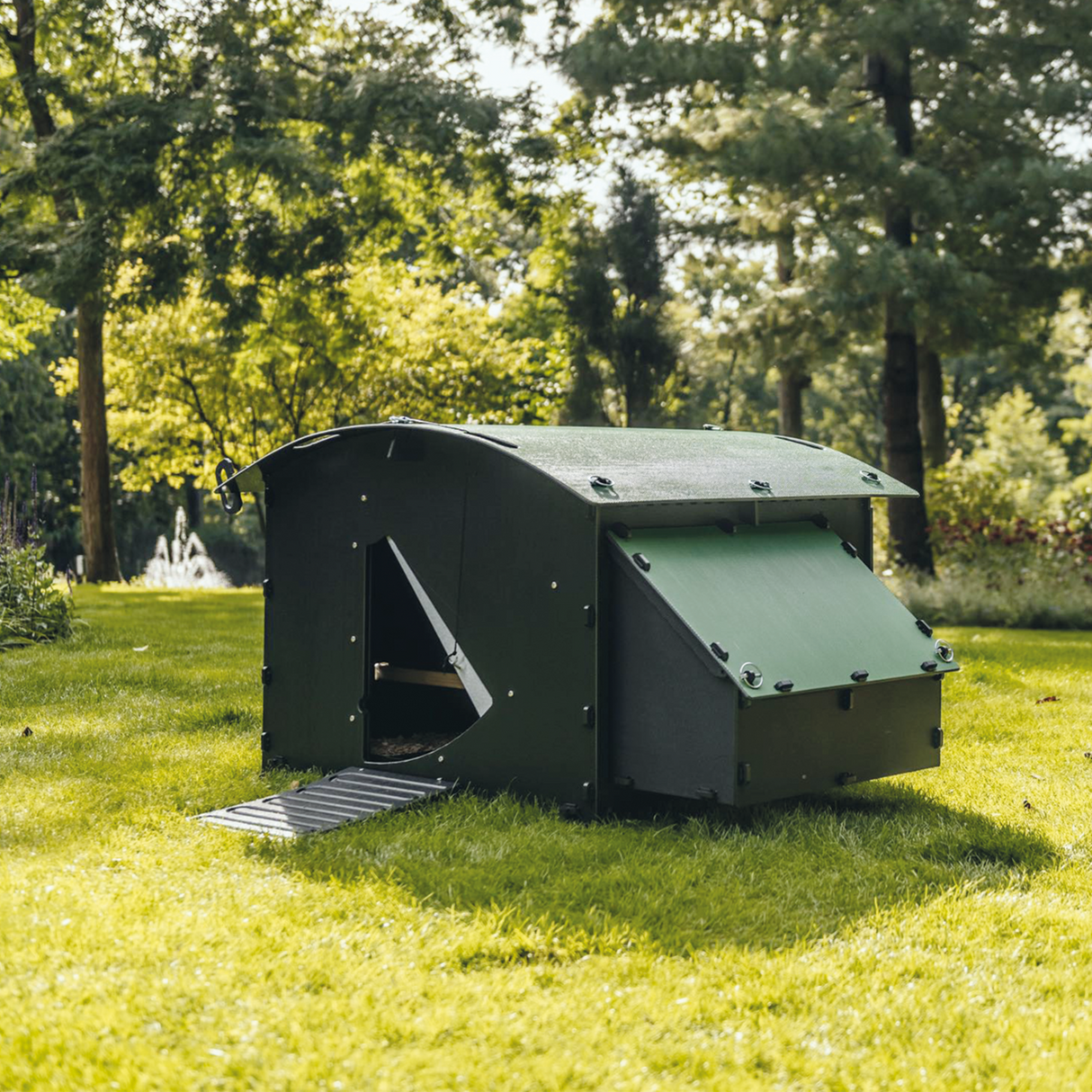 Hatching Time Nestera. Large coop can be seen in image in grass. There are trees in the background. Coop has door open with automatic coop opener. Ramp is leading up to opening for easy chicken access. Nesting box is on side connected to chicken coop, with lifting door for easy egg access. Roof of coop is slanted to prevent roosting.