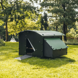 Hatching Time Nestera. Large coop can be seen in image in grass. There are trees in the background. Coop has door open with automatic coop opener. Ramp is leading up to opening for easy chicken access. Nesting box is on side connected to chicken coop, with lifting door for easy egg access. Roof of coop is slanted to prevent roosting.