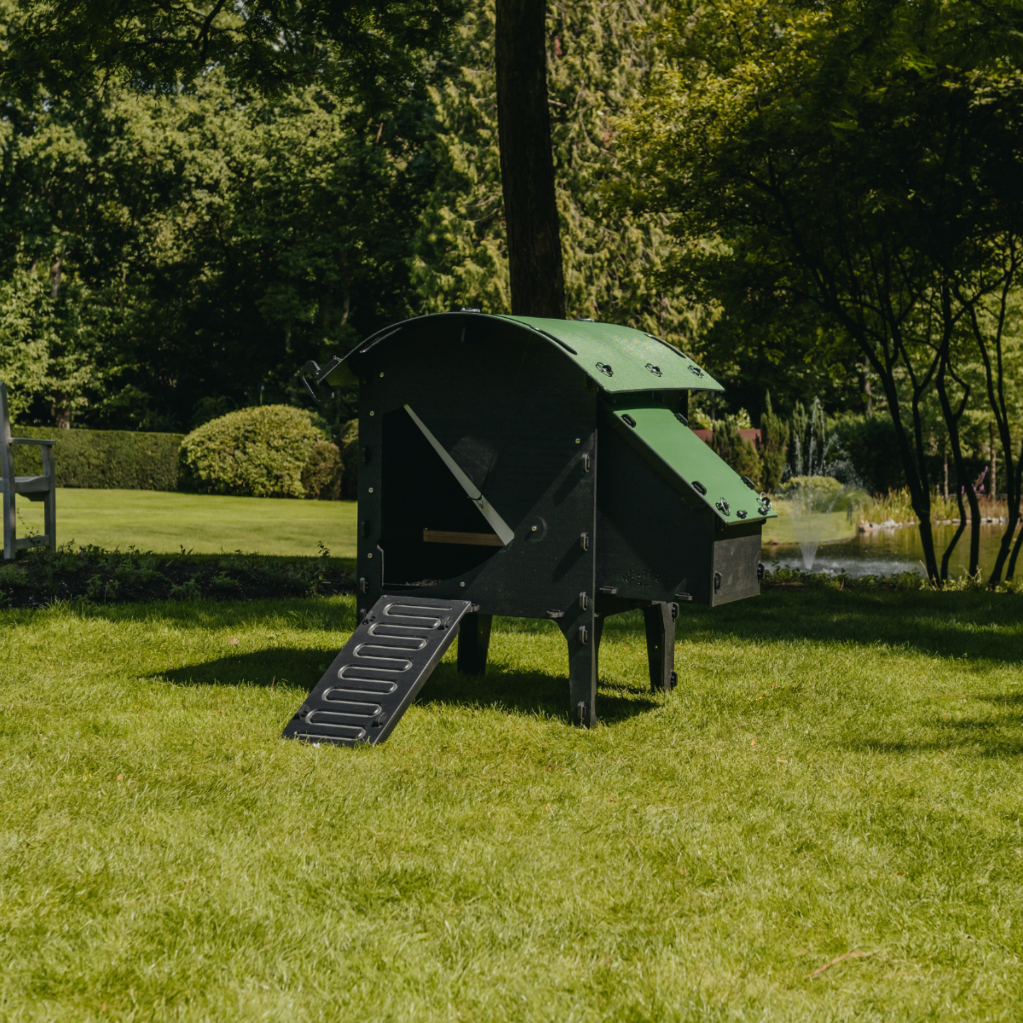 Hatching Time Nestera. Small Lodge coop on stand can be seen in image on grass. Coop has door open with automatic coop opener. Ramp is leading up to opening for easy chicken access. Nesting box is on side connected to chicken coop, with lifting door for easy egg access. Roof of coop is slanted to prevent roosting.