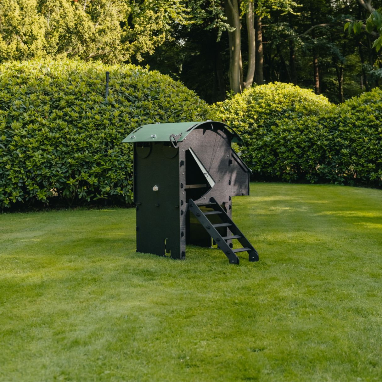 Hatching Time Nestera. Small Raised coop can be seen in image on grass. Coop has door open with automatic coop opener. Ramp is leading up to opening for easy chicken access. Nesting box is on side connected to chicken coop, with lifting door for easy egg access. Roof of coop is slanted to prevent roosting.