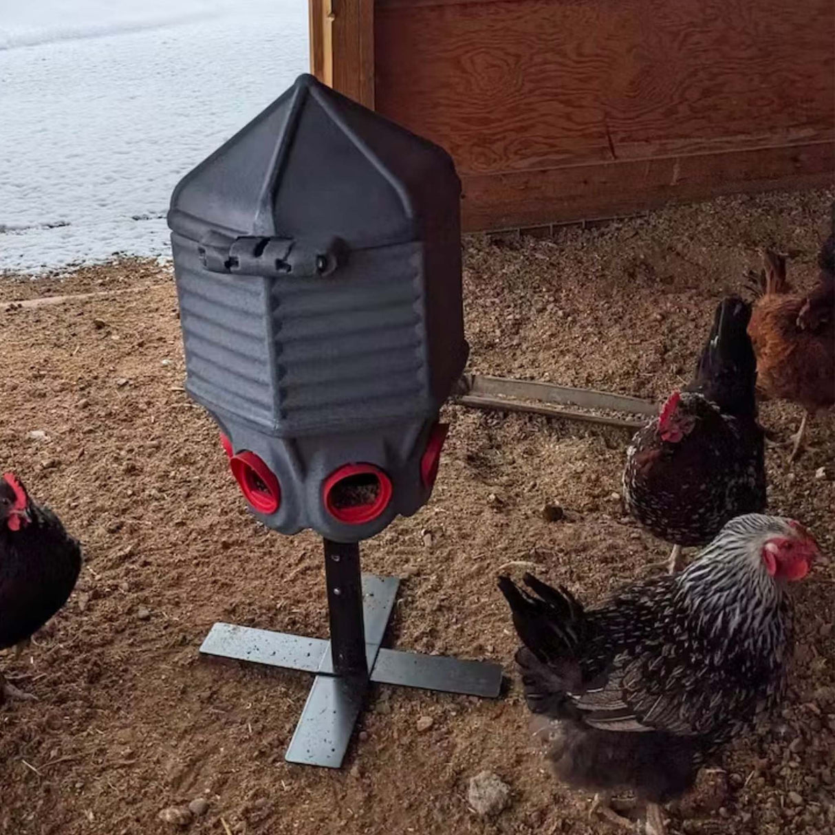 Gray bird feeder on a stand, surrounded by chickens outdoors.