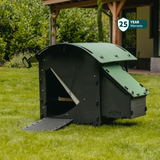 Hatching Time Nestera. Small house chicken coop can be seen on grass. Coop door is open with ramp leading to it for easy chicken access. Nesting box can be seen connected to side of chicken coop. 25 year warranty sticker can be seen in image.