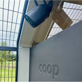 Close-up of a security camera on a Hatching Time Coop Camera branded structure with a metal fence background.